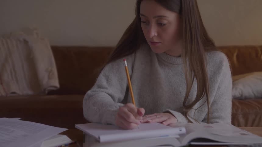 A woman in a gray sweater writes in a notebook at a cluttered coffee table