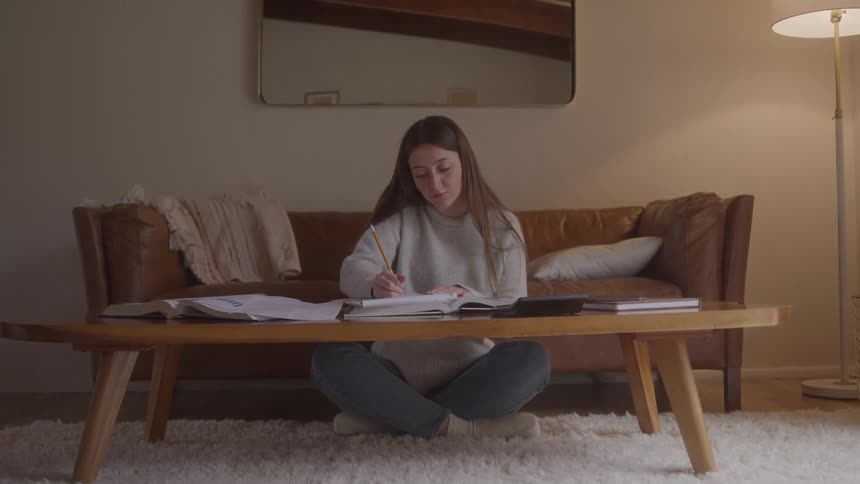 A young woman studies in her living room sitting on the floor and writing on a note placed on the table in front of her