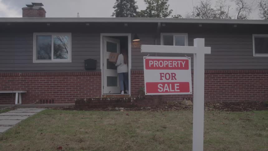 Woman enters the house carrying a box beside a property for sale sign