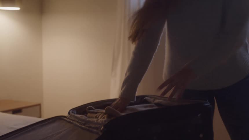 A female hurriedly packs an open suitcase in a bedroom