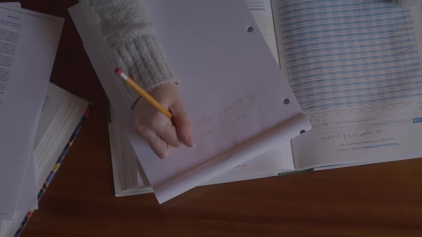 Woman writes with pencil on paper atop an open book surrounded by study materials