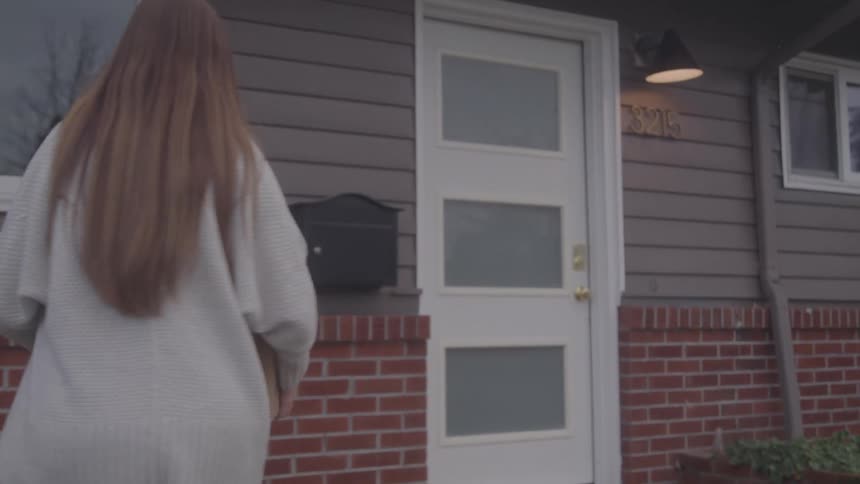 A female walks up to a house door carrying a large box then she opens the door