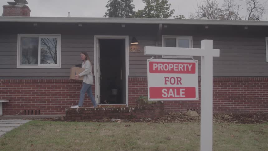 Teenager girl carrying a cardboard box out of the door of a house with a for sale sign in front outdoors during day time