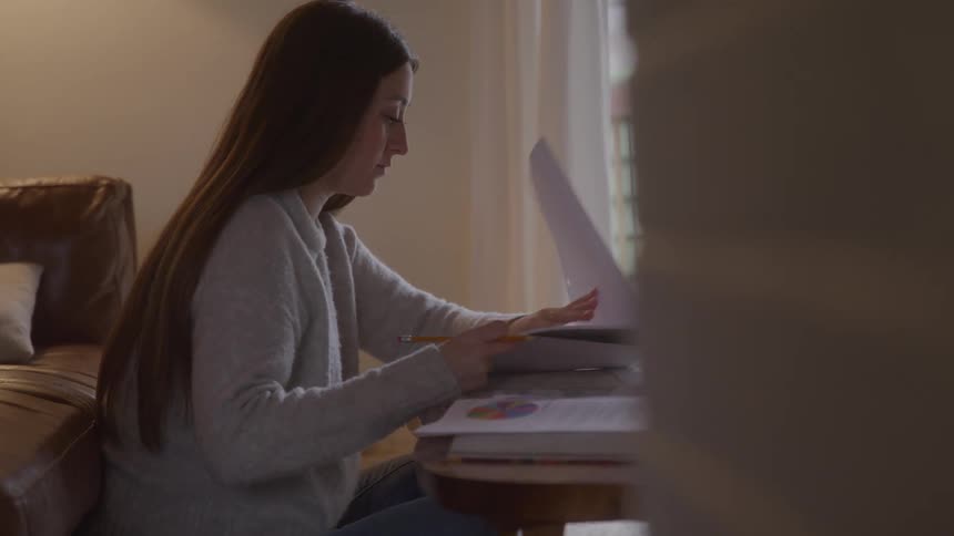 A woman sits at a table writing in a notepad on a table with books and papers
