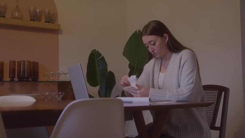A woman seated at a wooden table with an open laptop holding writing instruments and papers surrounded by domestic furnishings