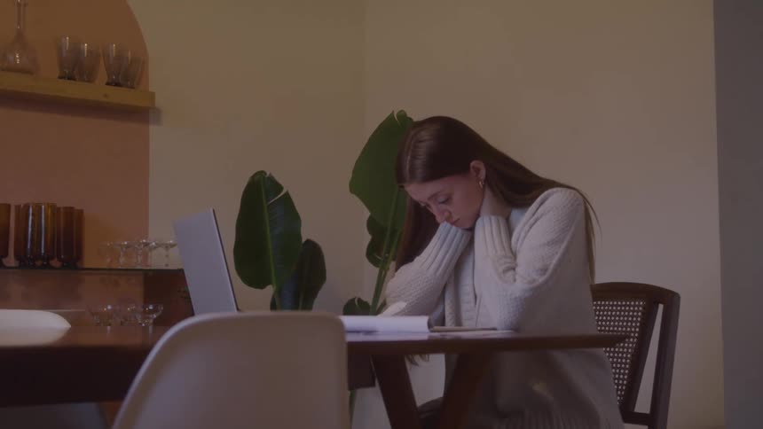Woman sitting at the table rests her head in her hands with a tired look in front of the laptop