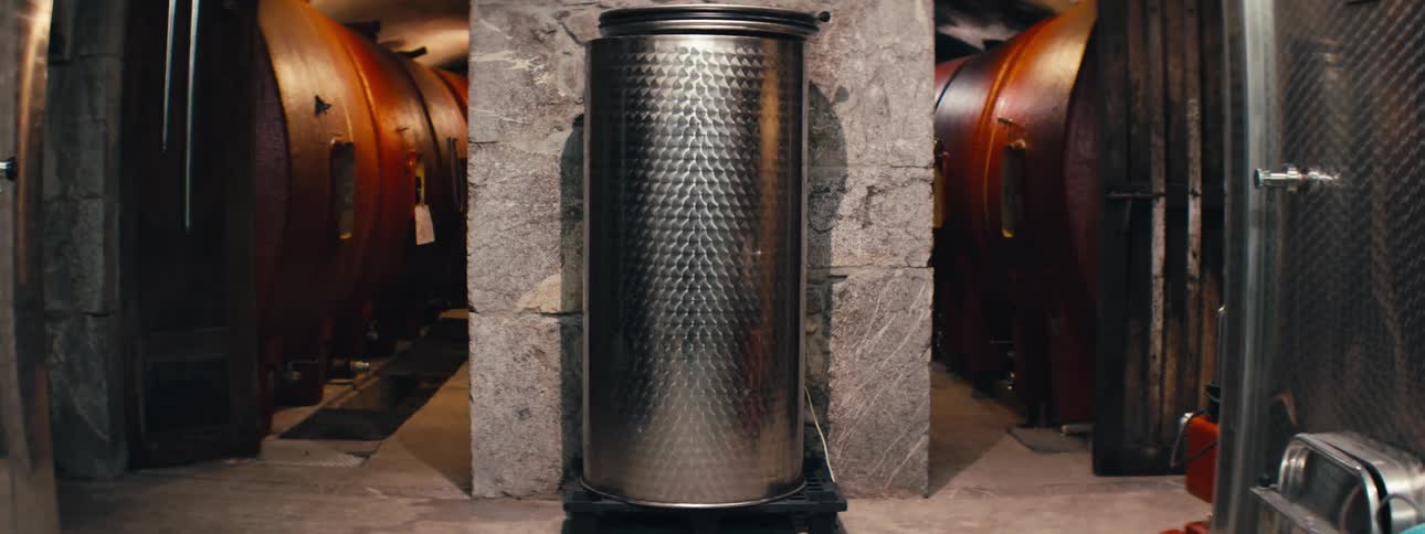Large metal tank stands in a cellar surrounded by wooden barrels
