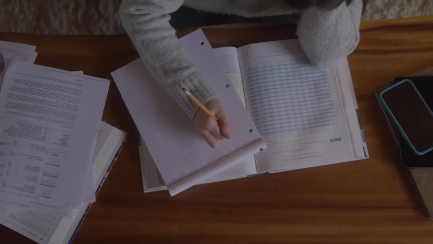 A student works on homework at a desk writing on paper placed over a textbook amidst scattered papers and a phone