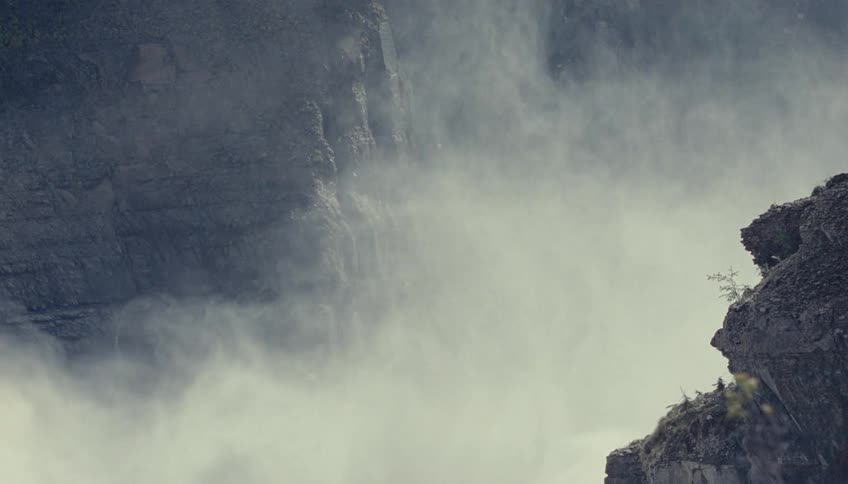 A wide lowangle view shows the spray and mist rising from a powerful waterfall between dark sheer rock cliffs in a natural outdoor environment