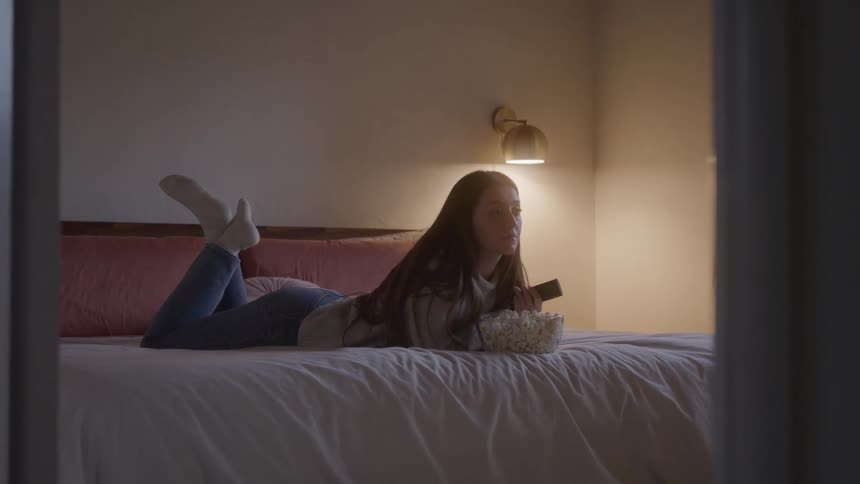 A young woman in jeans and socks lies on her bed eating popcorn watching tv and switching channels with a remote