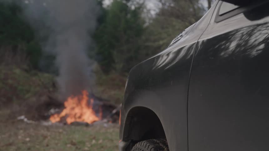 Dark vehicle parked in front of fire with thick smoke in a wooded outdoor area