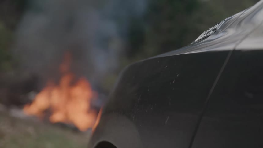 A car is parked on the roadside with a bonfire burning in front of it at midday
