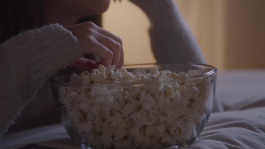 The woman is laying in bed eating popcorn