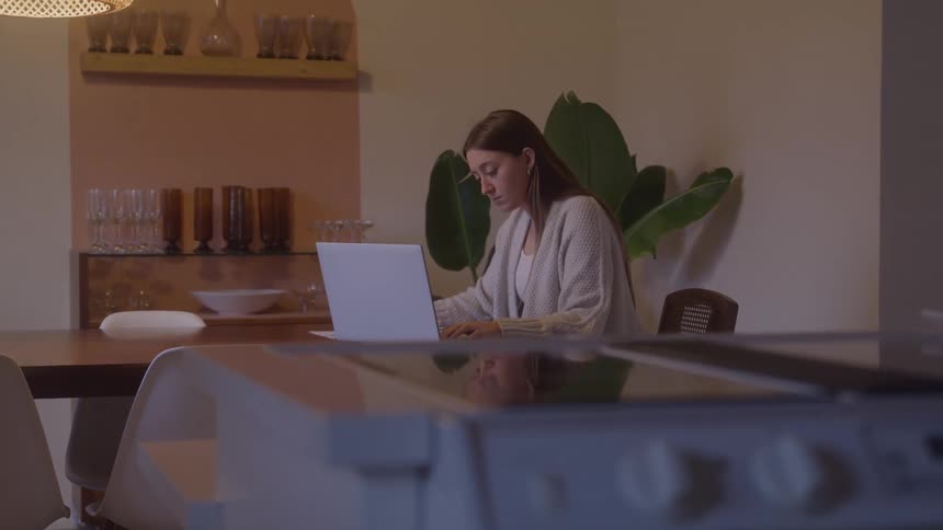 Woman works focused on a laptop in a cozy softly lit kitchen