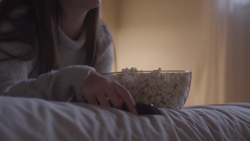 Person holds a remote control beside a bowl of popcorn on the bed