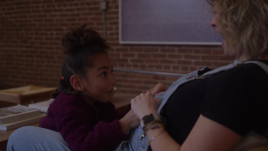 Pregnant mother seated interacts with daughter in indoor setting with brick wall