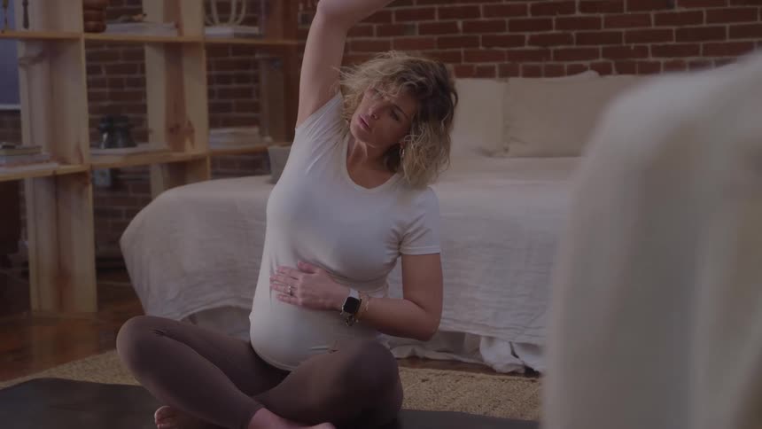 Pregnant woman performs side stretch seated on yoga mat inside bedroom