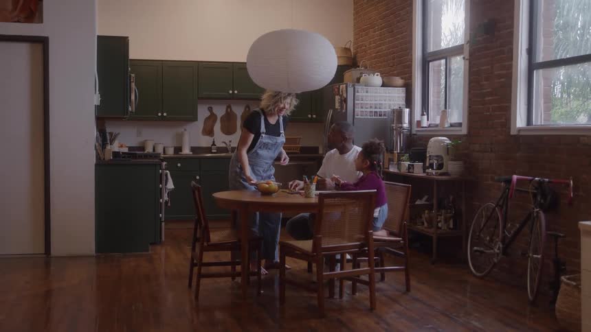 Mother brings a plate of food to the dining table to sit with her partner and daughter