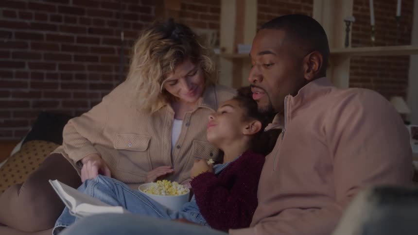 Girl eating popcorn while her father reads a book to her