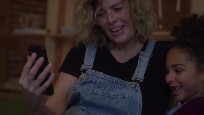 Two people sitting side by side look at phone while smiling in room with brick wall
