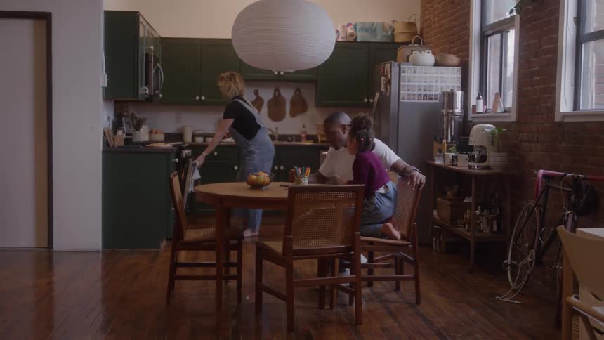 Mother preparing food in the kitchen while father and daughter discuss while sitting at the dining table indoors during daytime