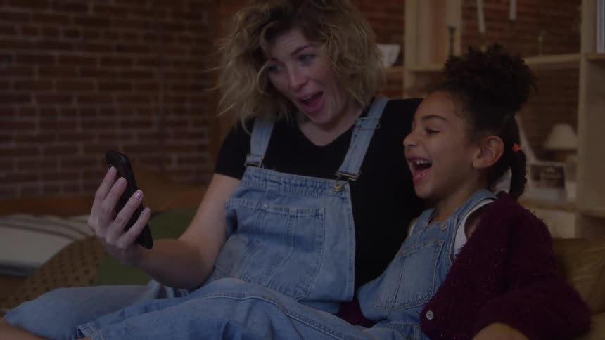 Two smiling people sitting on couch look at phone in room with brick wall