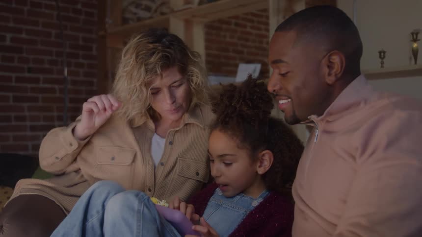 Father and mother sitting with their daughter on a couch in the living room while eating popcorn and watching media content from her tablet