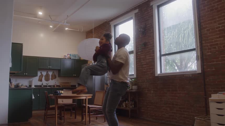 Father lifts daughter in affectionate gesture inside kitchen with brick wall and natural light