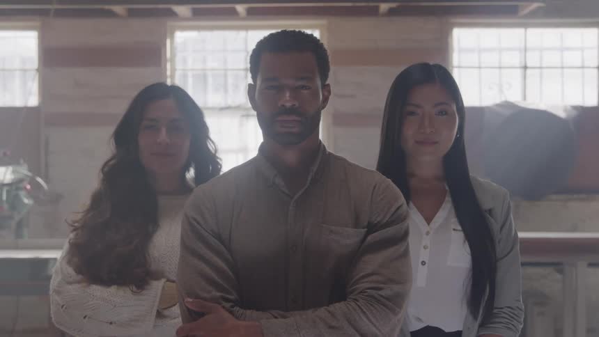 Three people standing indoors with large windows and wooden wall in the background