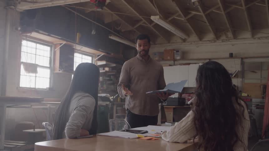A young man teaches two women in a factory at midday