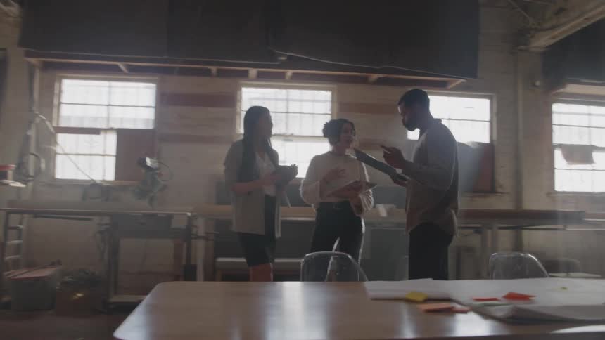 Three colleagues discuss blueprints and documents in a sunlit industrial workshop with large windows and tools visible in the background