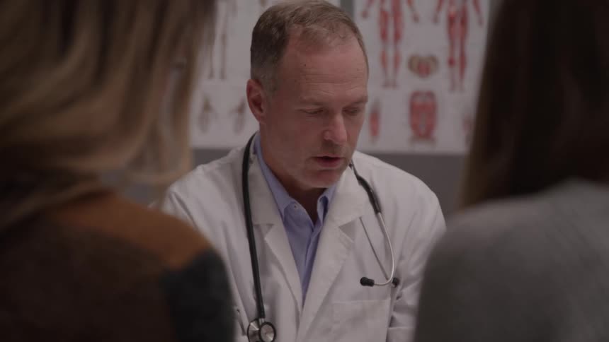 Doctor in white coat speaks with two patients in a clinical setting with anatomical charts in the background