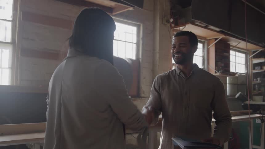 Man and woman shake hands in sunlit studio with large windows and work materials in background