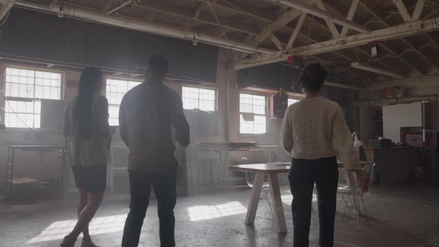 Three people one man and two women in smart casual business attire walk towards a table in a large dusty warehouse office space with exposed wooden rafters and bright windows
