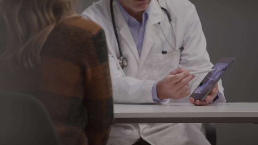Medical consultation with doctor pointing to information on tablet for patient in plaid sweater in a professional clinical setting