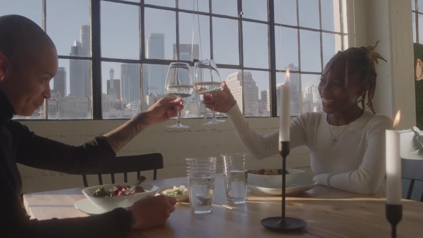 Couple toasts with wine at a decorated table in front of windows overlooking the city