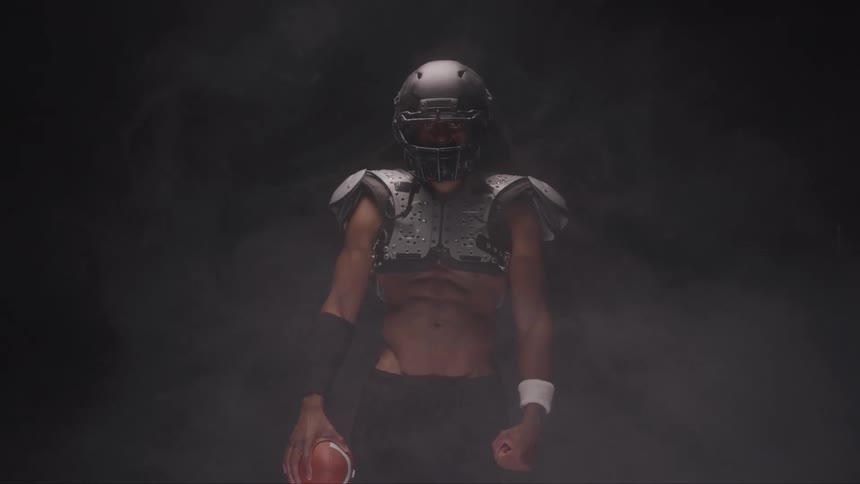 American football player in black helmet holds ball in dark smoky setting