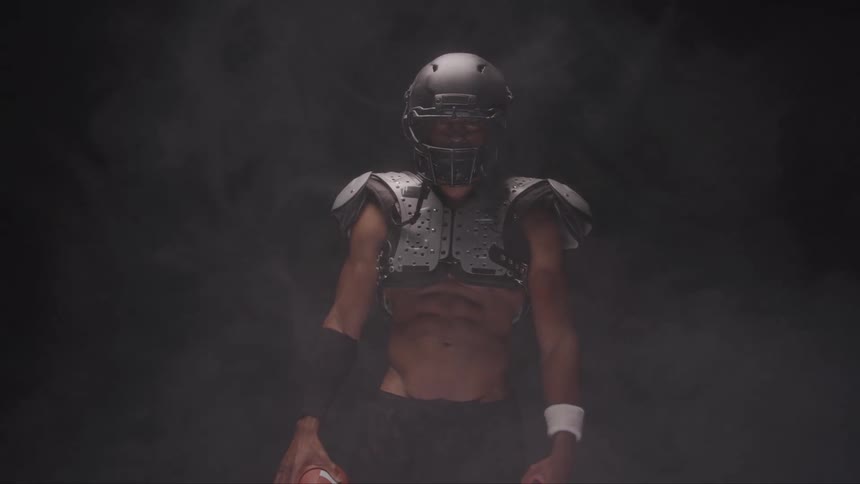 A muscular football player wearing a dark helmet and shoulder pads with his torso exposed stands holding a football in a dark smoky atmosphere with dramatic lighting