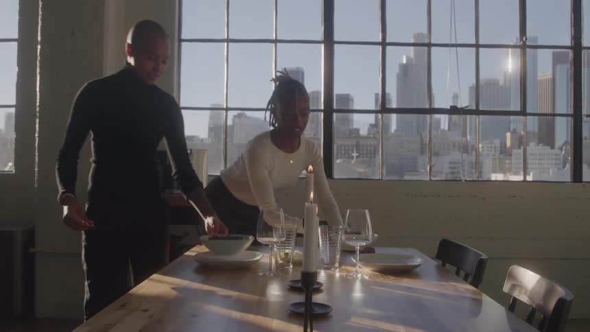 A young black couple arranging plates of food on a dining table for an indoor date during daytime