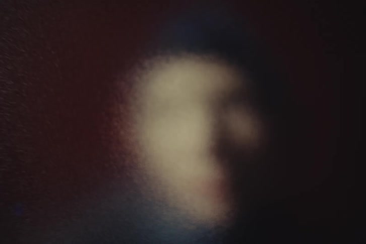 Persons face appears blurred behind textured glass in a dark setting