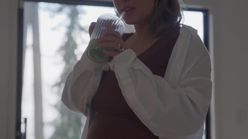 A pregnant woman stands indoors at midday drinking from a cup with a straw