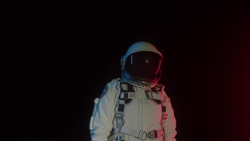 An astronaut in a white spacesuit stands against a completely black background illuminated by dramatic red lighting