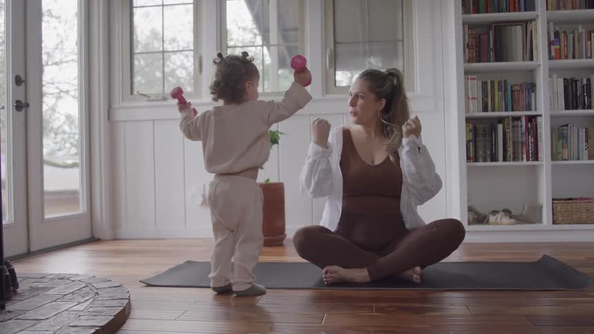 A pregnant woman wearing a brown workout outfit sits crosslegged on a yoga mat talking to a small child who stands facing away mimicking her with pink toy dumbbells raised overhead in a bright room with large windows and a bookshelf