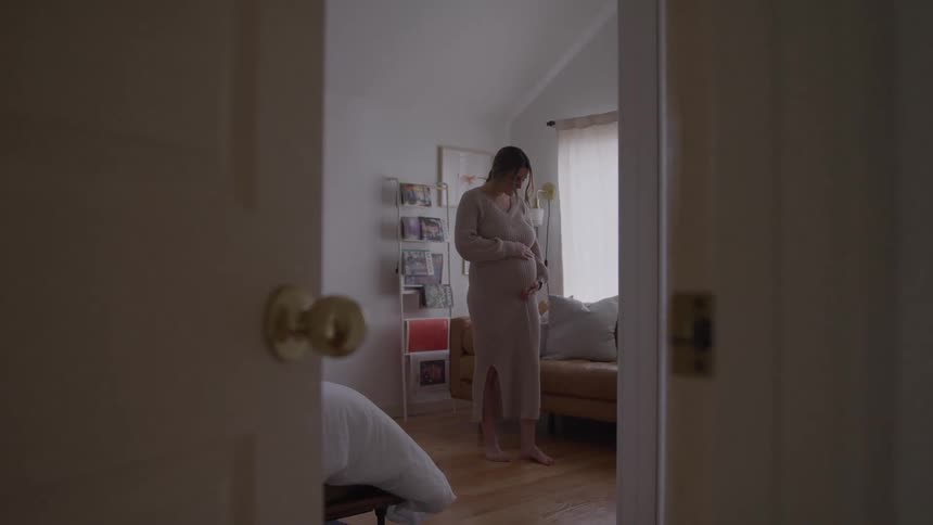 Woman standing in intimate setting touching belly in moment of contemplation