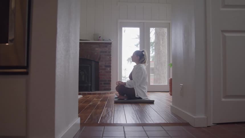 Mediumlong view framed by two walls of a woman sitting on a cushion with her legs crossed on a wooden floor in front of a fireplace and a glass exterior door