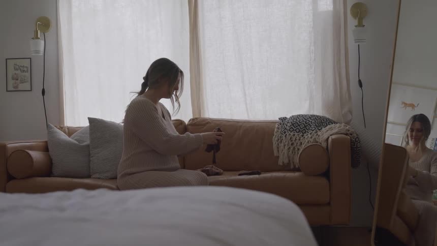 This shot is a static mediumwide view inside a modern bright bedroom with large windows likely in the morninga woman in a lightcolored outfit is sitting on a tan leather sofa folding a pair of dark socks while her reflection is clearly