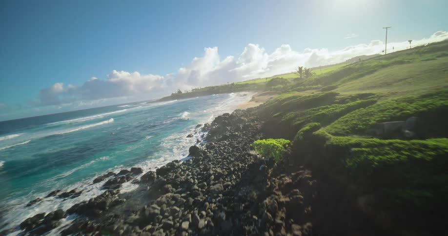 Ocean waves crash against a rocky shore under a blue sky with sunlit clouds
