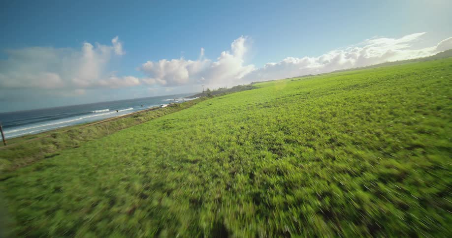 An aerial wide view sweeps down a steep green hillside toward a coastal highway running alongside the blue ocean with breaking waves under a bright blue sky with white clouds