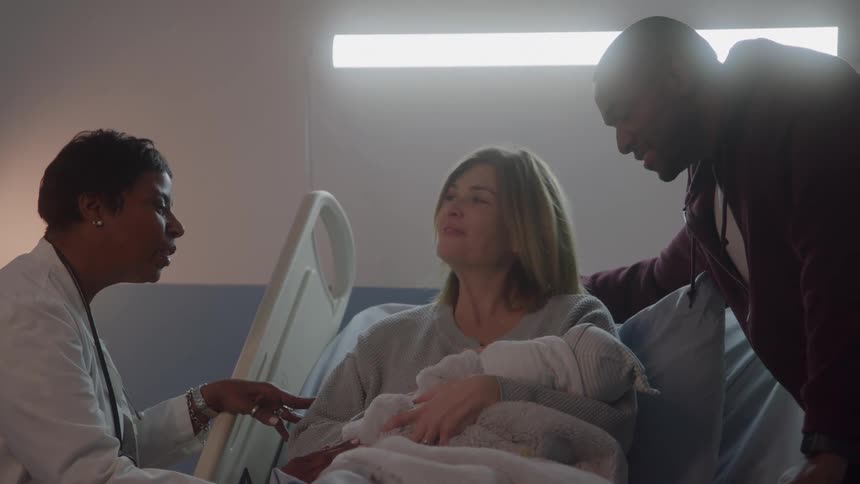 A woman lies in a hospital bed holding her newborn baby with a man by her side and a female doctor on the other side