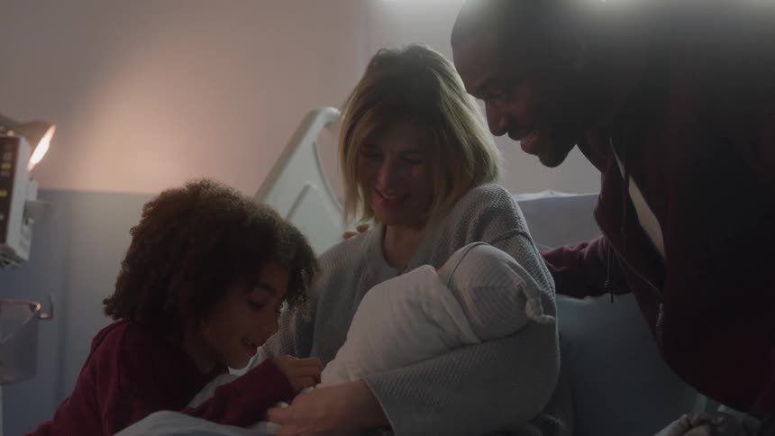 A family of four are in a hospital room rejoicing over the newborn the mother in the bed is holding